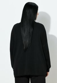 Frau mit langen, glatten schwarzen Haaren, die einen lockeren schwarzen Pullover und schwarze Hosen trägt, steht vor einer schlichten weißen Wand und ist von hinten zu sehen.