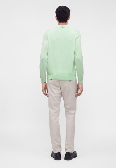Homme portant une chemise à manches longues vert clair et un pantalon beige, debout de dos face au spectateur sur un fond clair uni.