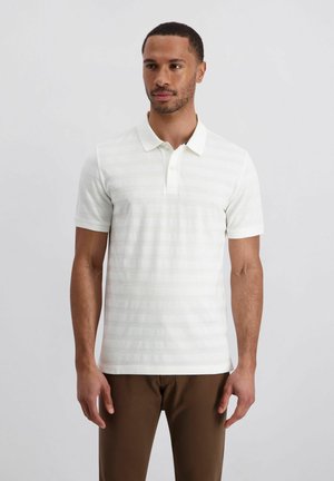 Homme portant un polo à manches courtes blanc avec des rayures subtiles et un pantalon marron, debout devant un fond clair uni.