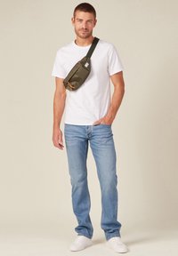 Riñonera verde oliva con detalles en naranja, llevada sobre una camiseta blanca y unos vaqueros azules. El modelo lleva zapatillas blancas y está de pie sobre un fondo neutro.