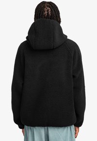 Chaqueta de forro polar negra con capucha, con un tejido texturizado, corte relajado y mangas raglán. Vista desde la parte de atrás, combinada con pantalones de color azul claro.