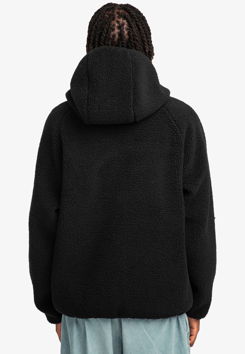 Giacca in pile nera con cappuccio, caratterizzata da un tessuto testurizzato, vestibilità comoda e maniche raglan. Vista da dietro, abbinata a pantaloni blu chiaro.