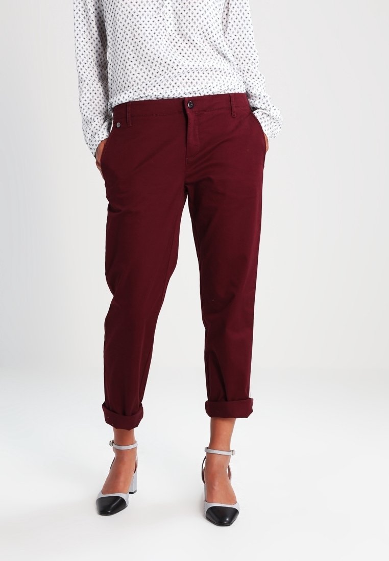 Femme portant un pantalon bordeaux retroussé, un chemisier blanc à motifs, et des talons à brides bicolores, debout devant un fond uni.
