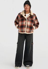 Vrouw met gevlochten haar, die een zwarte beanie, een rood geruit sherpa-jack, brede zwarte cargobroeken en zwart-witte sneakers draagt.