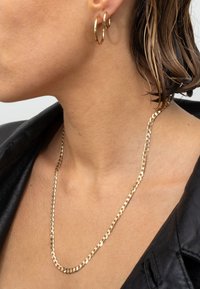 Goldfarbene Kettenhalskette mit einem glatten, gedrehten Design; kombiniert mit goldenen Creolen. Getragen über einer schwarzen Lederjacke.