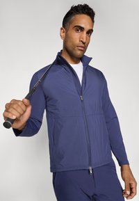Námořnická modrá golfová bunda vyrobená z lehkého, hladkého materiálu se zapínáním na zip, vysokým límcem a elastickým páskem; nošena s golfovým holem.