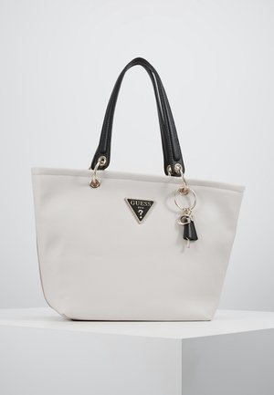 Sac fourre-tout blanc avec poignées noires, breloque anneau en métal et logo triangulaire Guess à l'avant, posé sur un socle blanc.