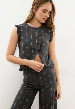 Femme portant un haut sans manches gris foncé boutonné et un pantalon assorti avec des motifs de feuilles bleu et gris, debout contre un mur blanc.