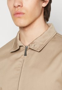 Chaqueta cremosa con cremallera, de textura suave, con cuello puntiagudo y cremallera de metal, que resalta líneas simples y un diseño minimalista.