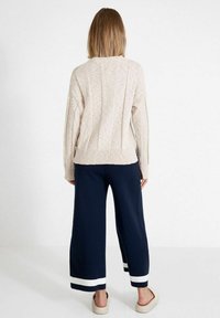 Beiger Strickpullover mit texturiertem Finish, kombiniert mit navyfarbener Hose in weiter Beinform, die am Saum weiße Streifen hat. Klobige Schuhe.