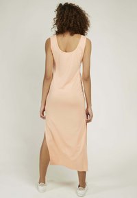 Robe longue sans manches de couleur pêche avec un dos plongeant, présentant une fente sur le côté et une texture lisse et extensible. Portée avec des baskets blanches.