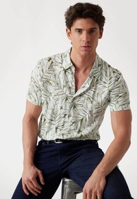 Camisa de manga corta en tela blanca con un patrón de hojas verdes, con un cierre de botones en la parte delantera y un diseño de cuello abierto. Se lleva con jeans de color azul oscuro.
