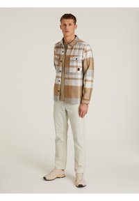 Camisa de cuadros en beige, blanco y gris con dos bolsillos en el pecho. Combinada con pantalones de colores claros y zapatillas beige. Textura de tela suave.