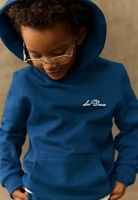 Παιδί με σγουρά μαλλιά που φορά χρυσά γυαλιά και μία μπλε κουκούλα με κεντημένο το "Les Deux", κοιτάζει κάτω με τα χέρια στις τσέπες.