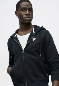 Tmavomodrá mikina na zips s predným vreckom, bielou vnútornou podšívkou a malým červeným trojuholníkovým logom na ľavej strane hrudníka.