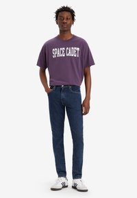 Camiseta de algodón morada con estampado de "SPACE CADET", combinada con jeans oscuros y zapatillas blancas con rayas negras. Corte casual.