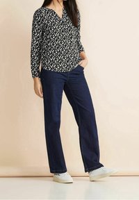 Schwarz-weiße gemusterte Bluse mit V-Ausschnitt, kombiniert mit dunkelblauen geraden Jeans und weißen Sneakers.