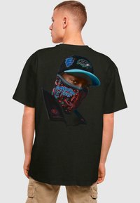 Svart T-shirt med ett grafiskt tryck av en person som bär en mönstrad mask och keps. Designen inkluderar livliga färger och logotyper.