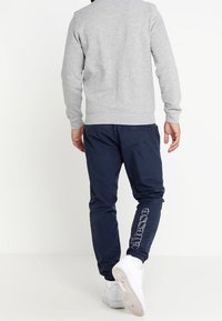 Grauer Sweatshirt mit Rundhalsausschnitt und gerippten Bündchen, kombiniert mit marineblauen Jogginghosen, die ein Logo auf dem linken Bein tragen. Weiße Sneaker runden das Outfit ab.