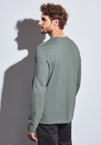 Camisa de manga larga con cuello redondo en algodón verde apagado, que cuenta con un corte relajado y dobladillo recto. Textura suave, sin patrones ni acentos visibles.