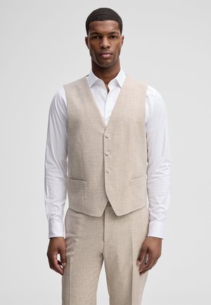 Mann trägt beigen Weste und Hose mit weißem Hemd, steht vor einem schlichten hellen Hintergrund.