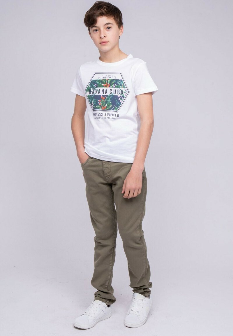 T-shirt blanche avec graphique tropical et texte, associée à un pantalon vert olive et des baskets blanches. Coupe décontractée, manches courtes, fond uni.