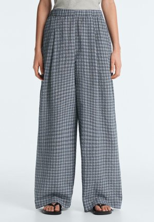 Kobieta ubrana w luźne szare spodnie w kratę z szerokimi nogawkami i czarne sandały, stoi na tle jednolitego jasnego tła.