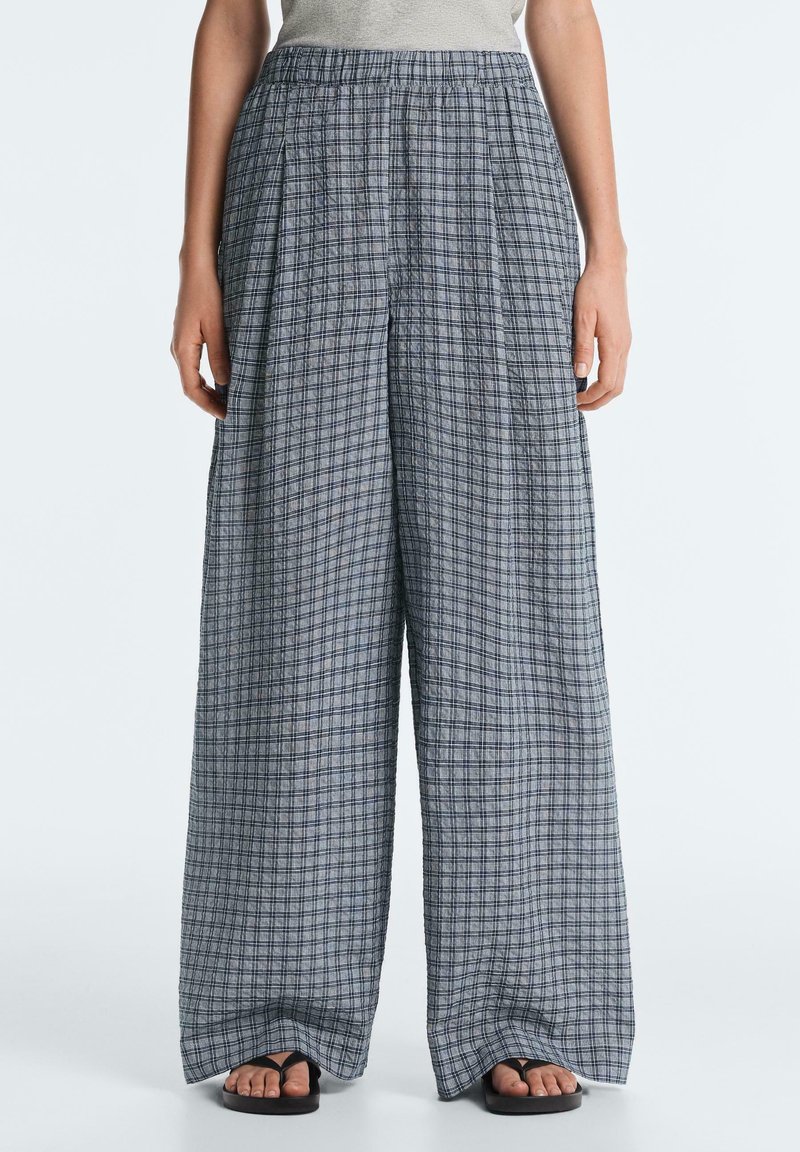 Femme portant un pantalon ample gris à carreaux à jambes larges et des sandales noires, debout devant un fond clair uni.