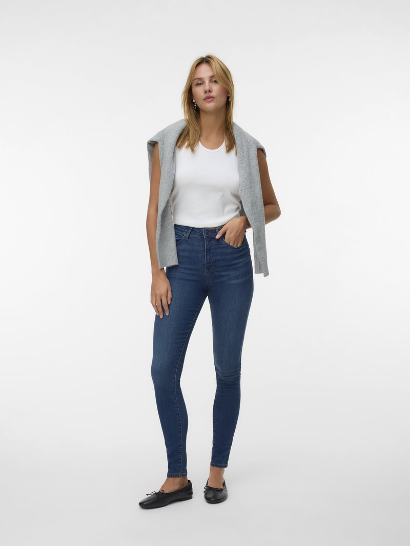 Graue ärmellose Strickjacke über einem weißen Tanktop, kombiniert mit blauen Skinny Jeans und schwarzen Ballerinas. Glatte Textur und taillierte Passform.