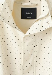 Veste blanche à pois noirs, fermeture éclair argentée partiellement ouverte, bouton-pression près du col, étiquette de la marque MNG à l'intérieur du col.