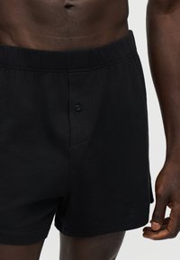 Personne à la peau foncée portant un boxer noir avec braguette boutonnée, montrant le ventre et la main droite près de la cuisse sur un fond uni.