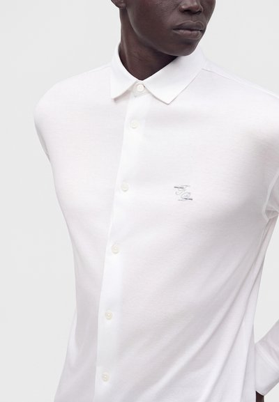 Homme portant une chemise blanche cintrée à boutons avec un col et un petit logo brodé sur la poitrine gauche, debout devant un fond uni.