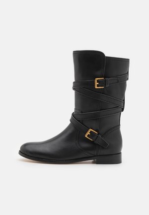 Czarny skórzany but do połowy łydki z dwoma ozdobnymi złotymi klamrami i owiniętymi wokół paskami, płaską podeszwą i zaokrąglonym noskiem.