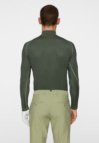 Långärmad topp i mörkgrön med geometriskt mönster. Åtsittande design, hög krage, kombinerad med ljusgröna byxor, visande bakvy.