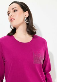 Magentové tričko s dlouhým rukávem, které má strukturovanou kapsu s jemnými stříbrnými ozdobami. Měkký materiál, kulatý výstřih a volný střih.
