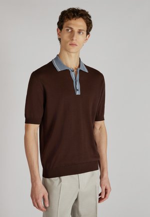 Mann in einem braunen kurzärmeligen Poloshirt mit blauem Kragen und hellgrauen Hosen, der vor einem schlichten Hintergrund steht.