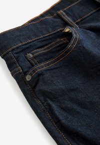 Tmavomodré džínsy z denimu v zábere zobrazujúce predný vrecko, mincovňové vrecko, oranžové švy a kovové nitky na bielom pozadí.