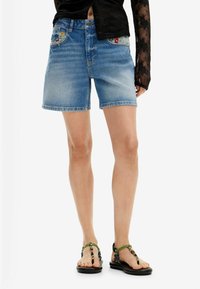 Pantalones cortos de mezclilla en azul claro con un diseño de corte recto, con bordado floral en los bolsillos y dobladillos deshilachados. Combinados con sandalias negras.