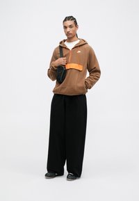 Hnedá fleece mikina s oranžovým akcentom a zipsom; spárovaná s voľnými čiernymi nohavicami a čiernymi topánkami; drží malú čiernu tašku.