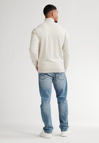Suéter de cuello alto de color crema con mangas largas, combinado con jeans de color azul claro y zapatillas blancas. Textura suave, diseño ajustado.