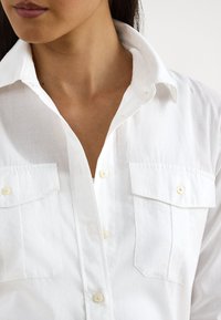 Camisa branca de botão com gola, dois bolsos com aba no peito e textura de tecido limpa. Os botões são claros e redondos.