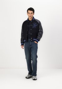 Veste varsity marine avec manches en cuir, col et poignets côtelés. Logo brodé à l'avant. Un jean bleu et des chaussures de sport grises complètent la tenue.