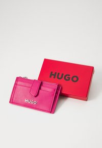 Roza usnjena denarnica z več režami in z zadrgo, ki je priložena rdeči škatli z logotipom "HUGO" v izrazitih črnih črkah.