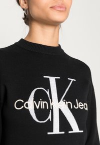 Černá pletená mikina s žebrovaným výstřihem a velkým grafickým potiskem v bílé a béžové barvě, na kterém je napsáno "Calvin Klein Jeans" a "CK".