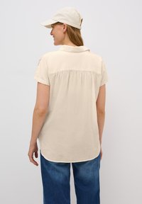 Beige kortebloemshirt met een ronde zoom en geplooide achterkant, gecombineerd met blauwe wijde spijkerbroek en een lichtgekleurde pet.