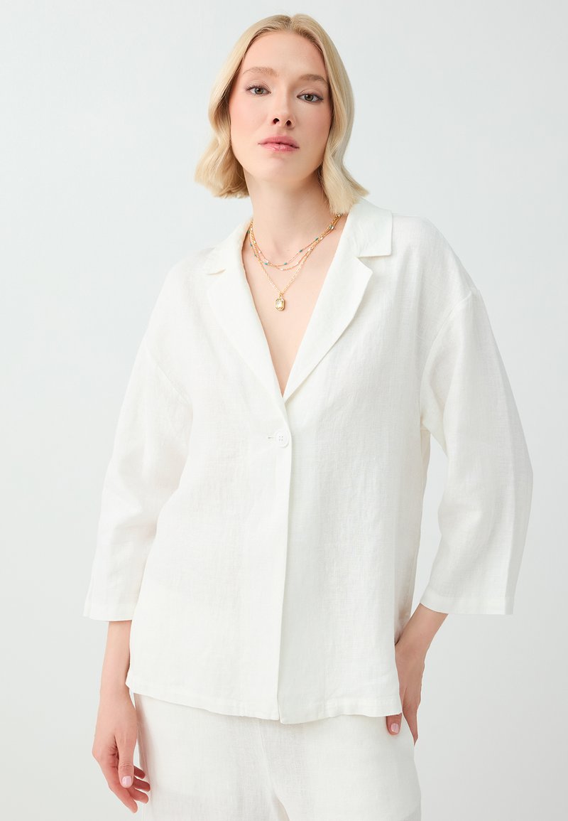 Femme blonde portant une chemise blanche ample boutonnée et un pantalon assorti, avec des colliers en or superposés sur un fond clair uni.