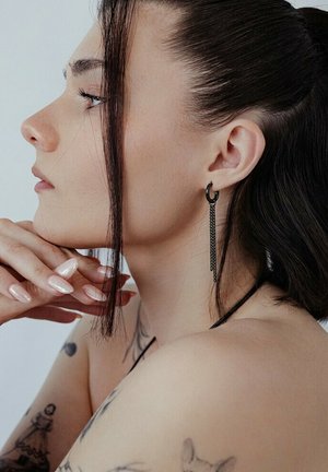 Woman in profile with dark hair, resting chin on hands, wearing long black chain earrings, and visible tattoos on shoulder and upper arm.
