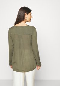 Olive green, long-sleeve blouse with a rounded neckline and back pleat detail. Lightweight, semi-sheer fabric, slightly curved hem.