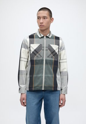 Junger Mann mit kurzen Haaren, der ein langärmliges Hemd mit Reißverschluss und großem grau-schwarz-weiß-braun kariertem Muster sowie blaue Jeans trägt.