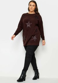 Bordeauxroter Strickpullover mit Paillettensternmustern, rundem Halsausschnitt und langen Ärmeln, kombiniert mit enganliegenden schwarzen Hosen und schwarzen Absatzstiefeln.